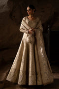 Woman in an ornate beige dress standing against a textured wall.