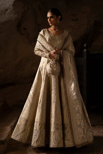 Woman in an ornate beige dress standing against a textured wall.