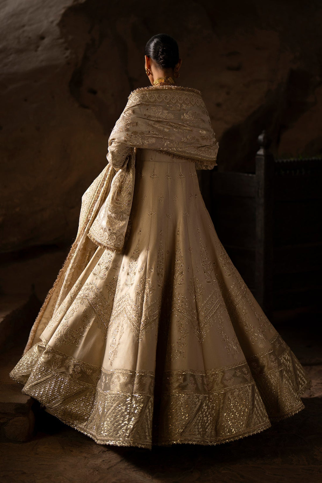 Woman in an ornate beige dress standing against a textured wall.